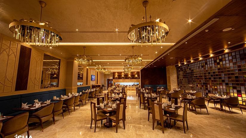 An elegant, warm-toned restaurant at The Gaurs Sarovar Premiere. Tables and chairs are set for dining. Decorative chandeliers illuminate the space. The room has a polished floor and patterned walls, creating a sophisticated atmosphere.