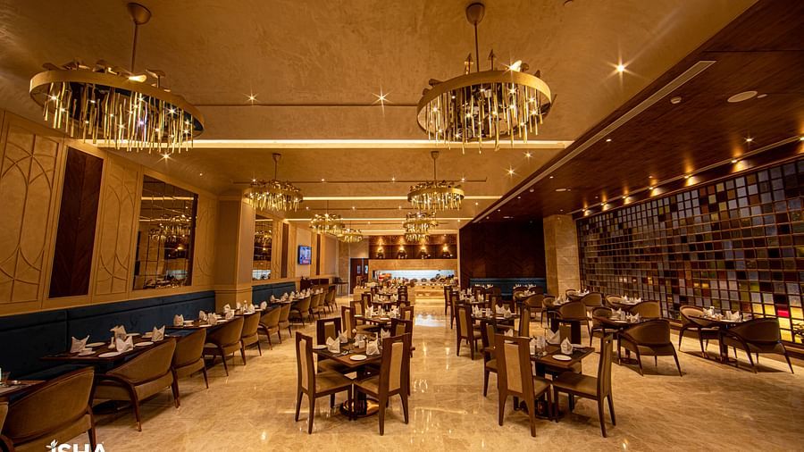 An elegant, warm-toned restaurant at The Gaurs Sarovar Premiere. Tables and chairs are set for dining. Decorative chandeliers illuminate the space. The room has a polished floor and patterned walls, creating a sophisticated atmosphere.