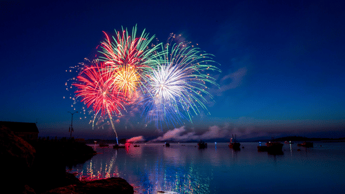 An overview of a lake in the night with fireworks in the sky, illuminated on the waters