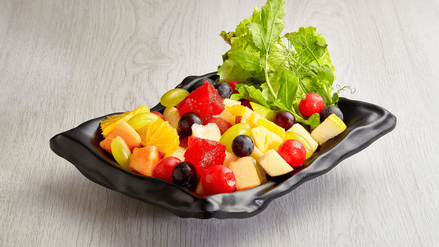 Black ruffled dish holding a citrus salad of orange segments, watermelon, grapes, papaya, pineapple, blueberries and lettuce leaves on wood.