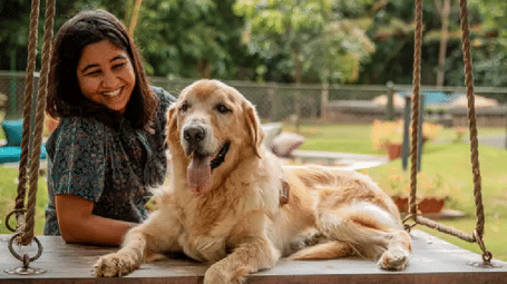 Guest sitting on the ground with a dog at the Karma Chalets outdoor area, grassy space with a fence in the background.