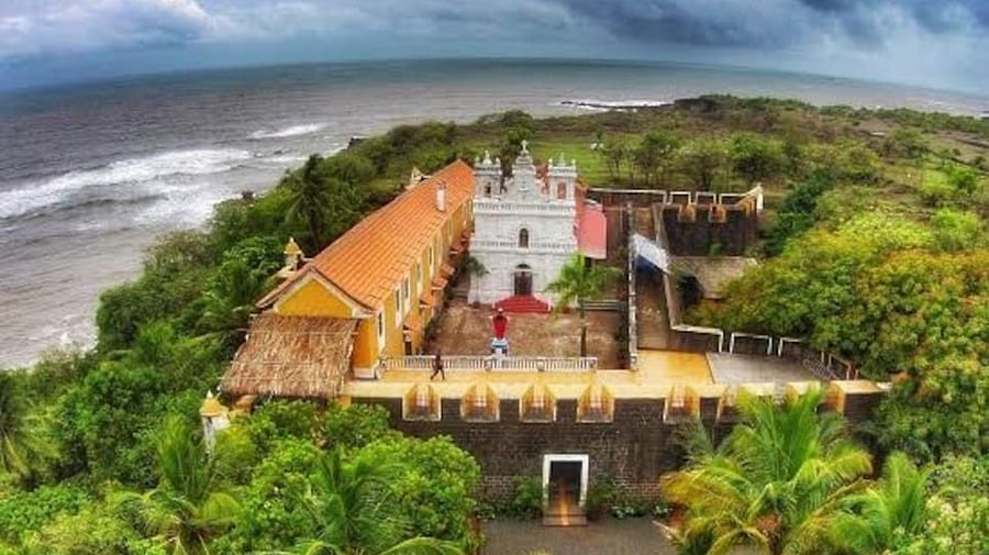 An aerial or high-angle view of Fort Tiracol, Goa, showing the white fort building with a red roof surrounded by lush green trees and overlooking the ocean under a dramatic, cloudy sky.