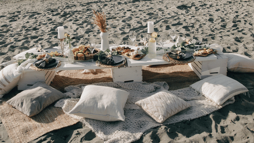 A rug kept on the beach with many items on it like pillows, food items, drinks and candles