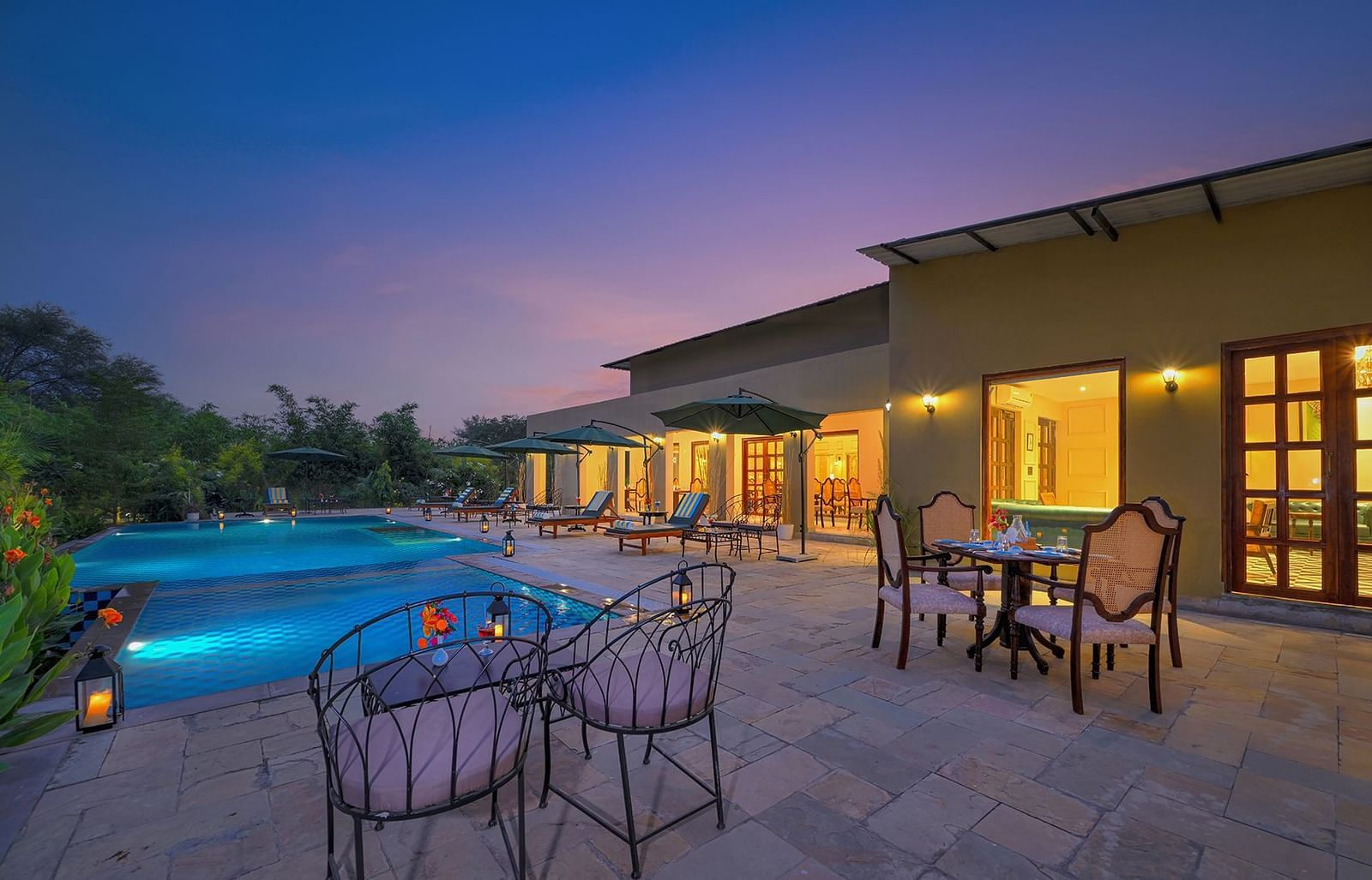 Poolside seating area with cushioned chairs and dining tables, surrounded by greenery and sun loungers under large umbrellas, illuminated by lanterns at dusk - Singhvilas, Ranthambore