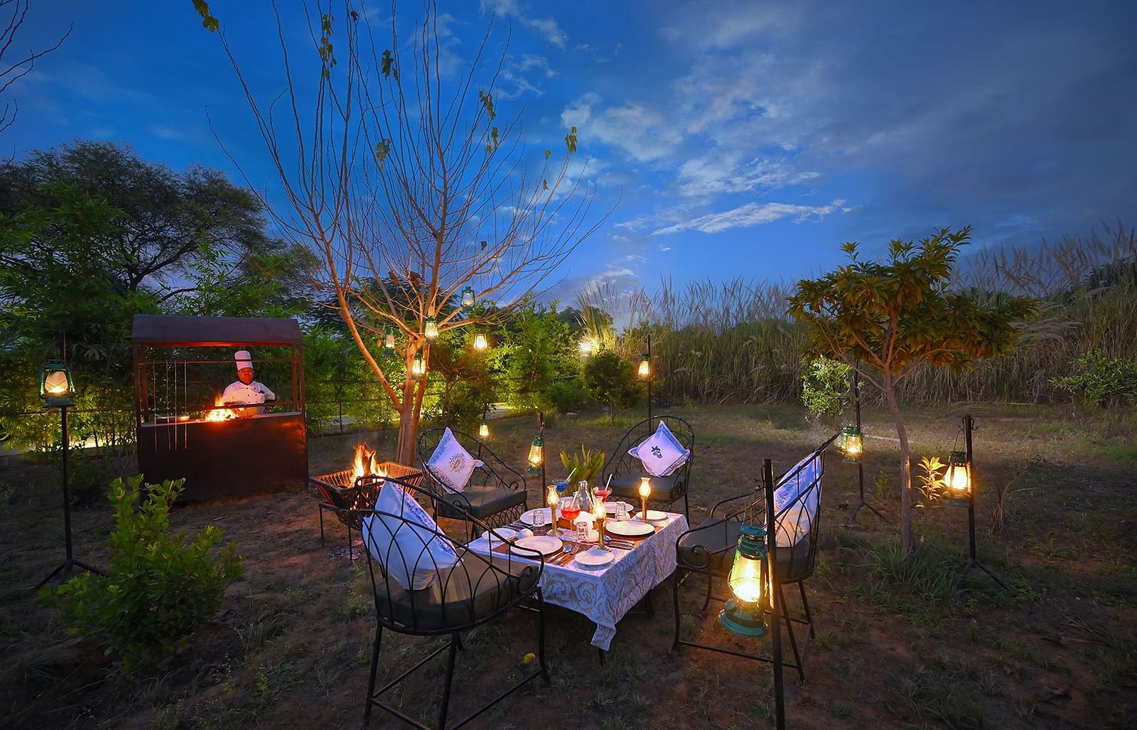 Private outdoor dining setup in a natural garden with cushioned chairs around a table adorned with lanterns and a live barbecue station, set against a serene twilight sky - Singhvilas, Ranthambore