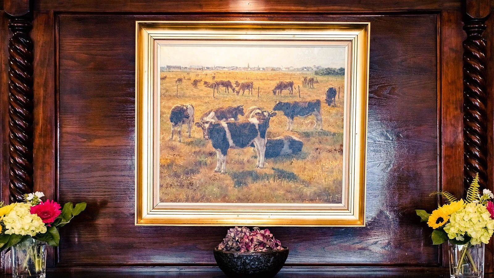 A framed painting of grazing cattle placed on a wooden mantel, decorated with  flowers and a small decorative bowl.- The Grand Hotel & Ballroom, McKinney