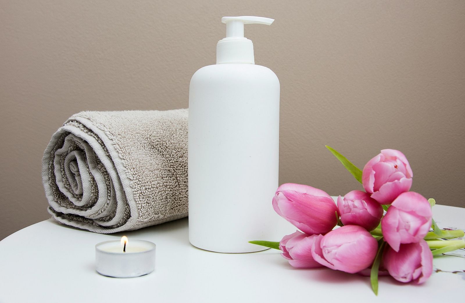A white pump bottle, folded towels, a lit candle, and pink tulips on a surface.