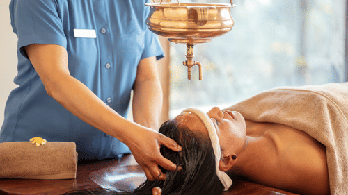 Woman receiving a traditional Ayurvedic head massage with warm oil being poured onto her forehead, administered by a therapist in a spa setting.  