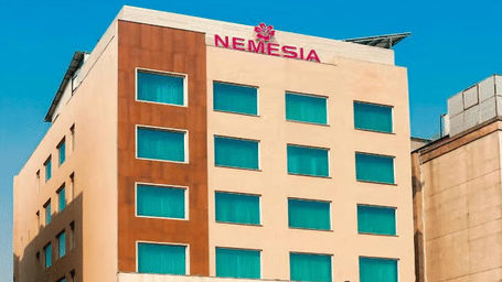 Daytime view of the Nemesia hotel building facade with teal-tinted windows, wooden accents, and palm trees in front.