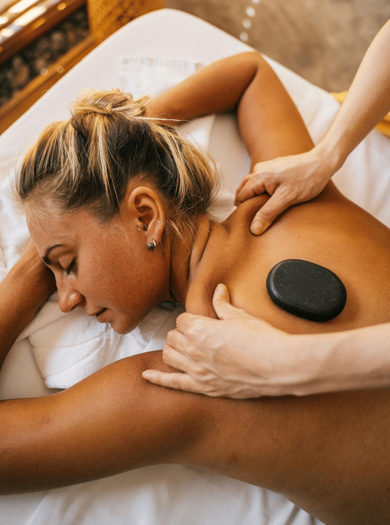 A woman receives a relaxing hot stone massage on her back, lying face down on a white spa bed.