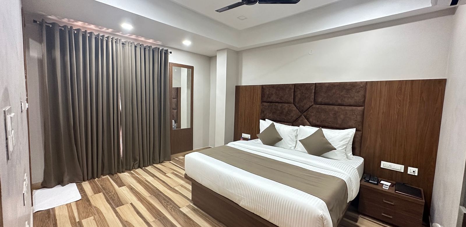 A room displays a double bed with white bedding, dark wooden headboard, and wooden flooring - Kankidham Lords Inn
