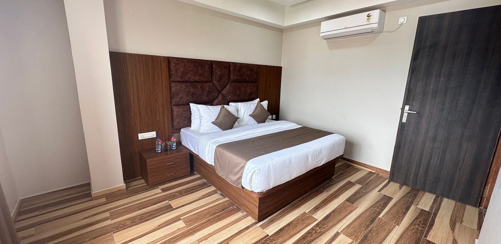 A bedroom includes a double bed with white and beige linens, dark wooden furniture, and a ceiling fan - Kankidham Lords Inn