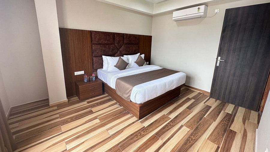 A bedroom contains a double bed with white bedding, a dark wooden headboard, a ceiling fan, and light walls - Kankidham Lords Inn