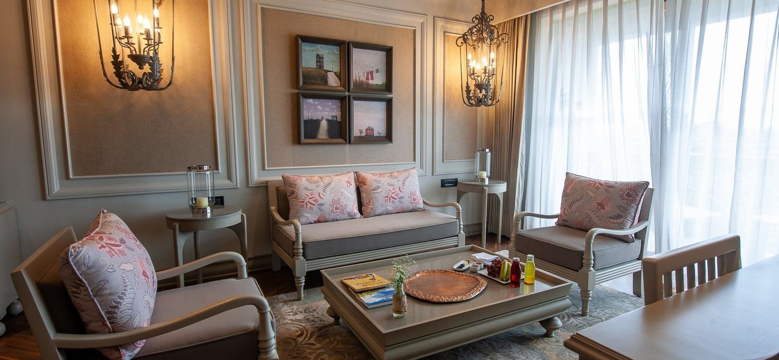The sophisticated living room of a suite at The Evren, Vagator, showcasing a seating arrangement with a sofa and two chairs around a coffee table. The room is decorated with framed art, a large rug, and two striking chandeliers.