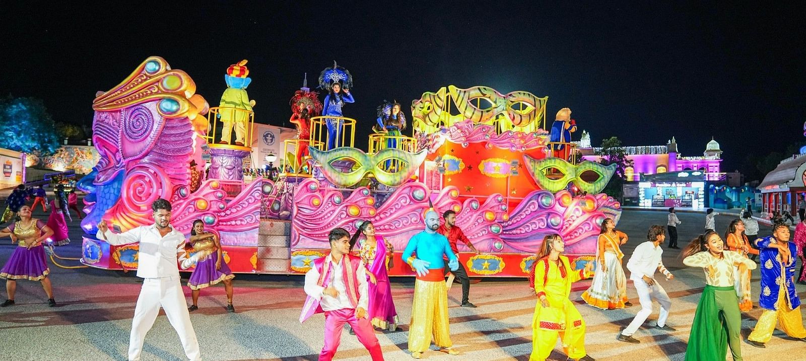 A vibrant parade with performers in colorful costumes dancing on a street at night. Brightly decorated floats with oversized masks and ornaments are in the background, creating a festive atmosphere.