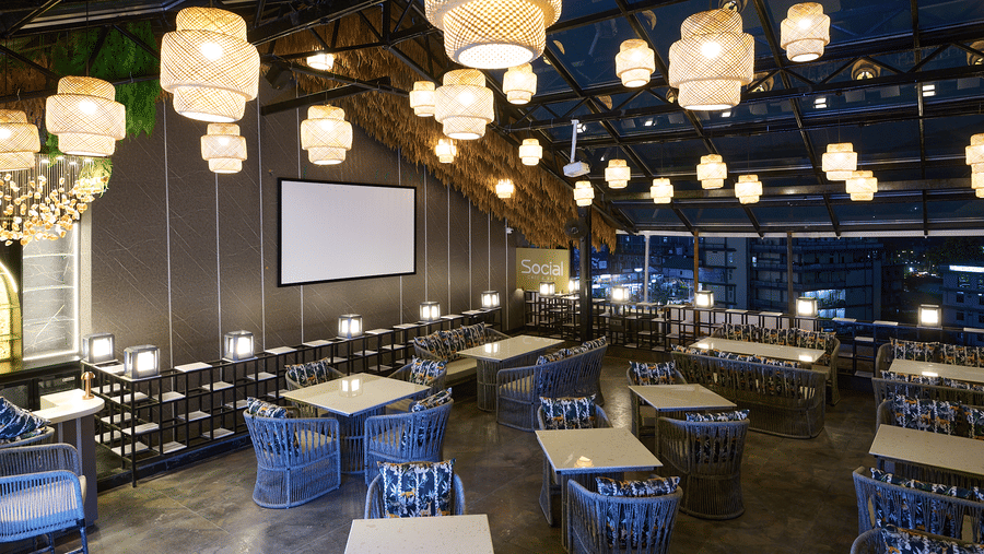 An indoor restaurant or dining area with blue chairs and tables, illuminated by multiple hanging lantern-style lights, and a projection screen in the background | Voyage Eastin Suites