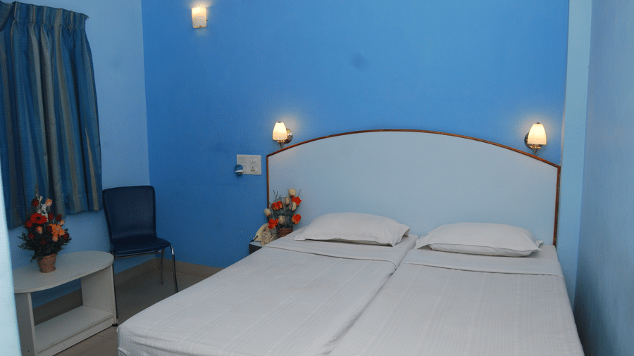 A clean hotel room setup featuring 2 single beds placed side by side against a bright blue wall with bedside lamps - Hotel Pandian