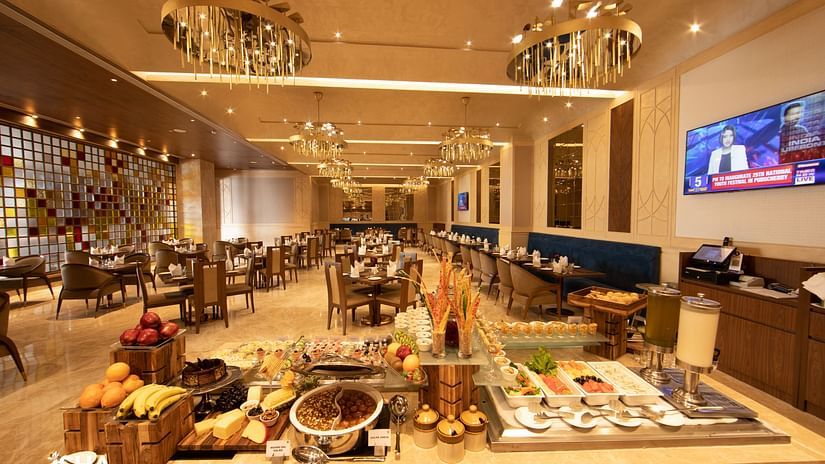 A brightly lit dining area at The Gaurs Sarovar Premiere. A buffet setup displays various food items. Tables and chairs are arranged for guests. Decorative chandeliers hang from the ceiling, and a television is visible.