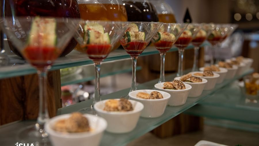A long display of small dessert cups and stemmed glasses filled with layered desserts, likely at a buffet. The desserts feature fruit, cream, and other colorful components. Bottles and other buffet items are visible in the blurred background.