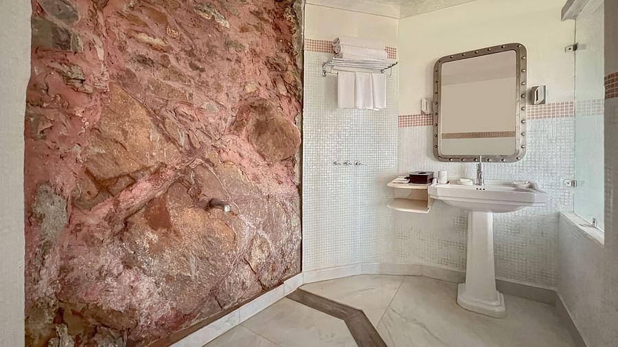 A bathroom with a shower area featuring exposed rock, a pedestal sink with a mirror above it, and towel racks at Tijara Fort-Palace - 19th Century, Alwar.