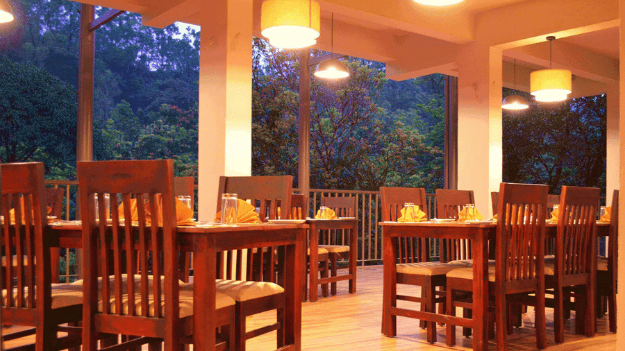A dining area with wooden chairs and warm lighting provides a cosy atmosphere with a view of the outdoors.- Abad Brookside Lakkidi, Wayanad