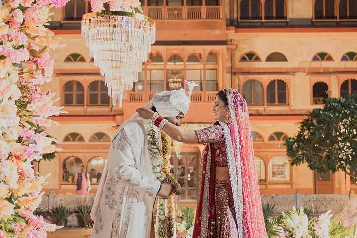 The bride places a garland on the groom under a floral canopy, with a grand palace backdrop illuminated by lighting. - Umaid Palace - A Lakeside Heritage Palace Resort
