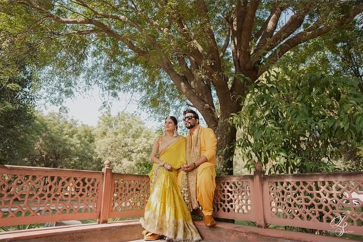 The couple poses under a large tree, holding hands and exuding elegance in a serene outdoor setting. Umaid Palace - A Lakeside Heritage Palace Resort