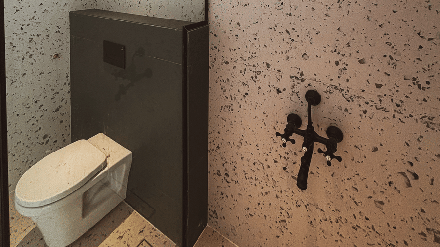 A black shower head and taps are mounted on a speckled wall next to a white toilet and a glass partition at Avanya Resorts N Retreat.
