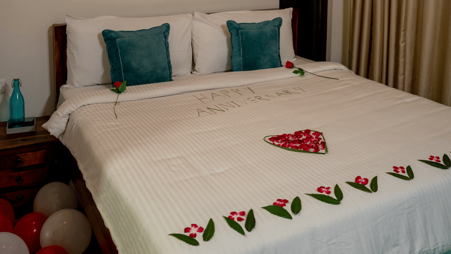 A bed with pillows and a runner sits in the Sun Terrace Room at BluSalzz Escapade Resort, Lansdowne. Decorative items are arranged on the bed and floor. A window with curtains is visible in the background.