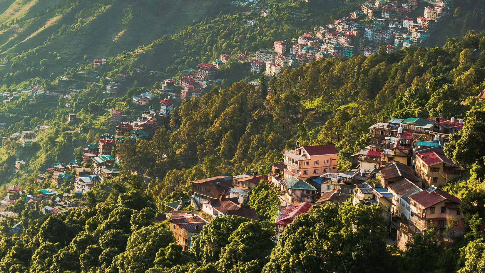An aerial view of a hillside village with colourful buildings nestled amidst dense green forest under bright sunlight.