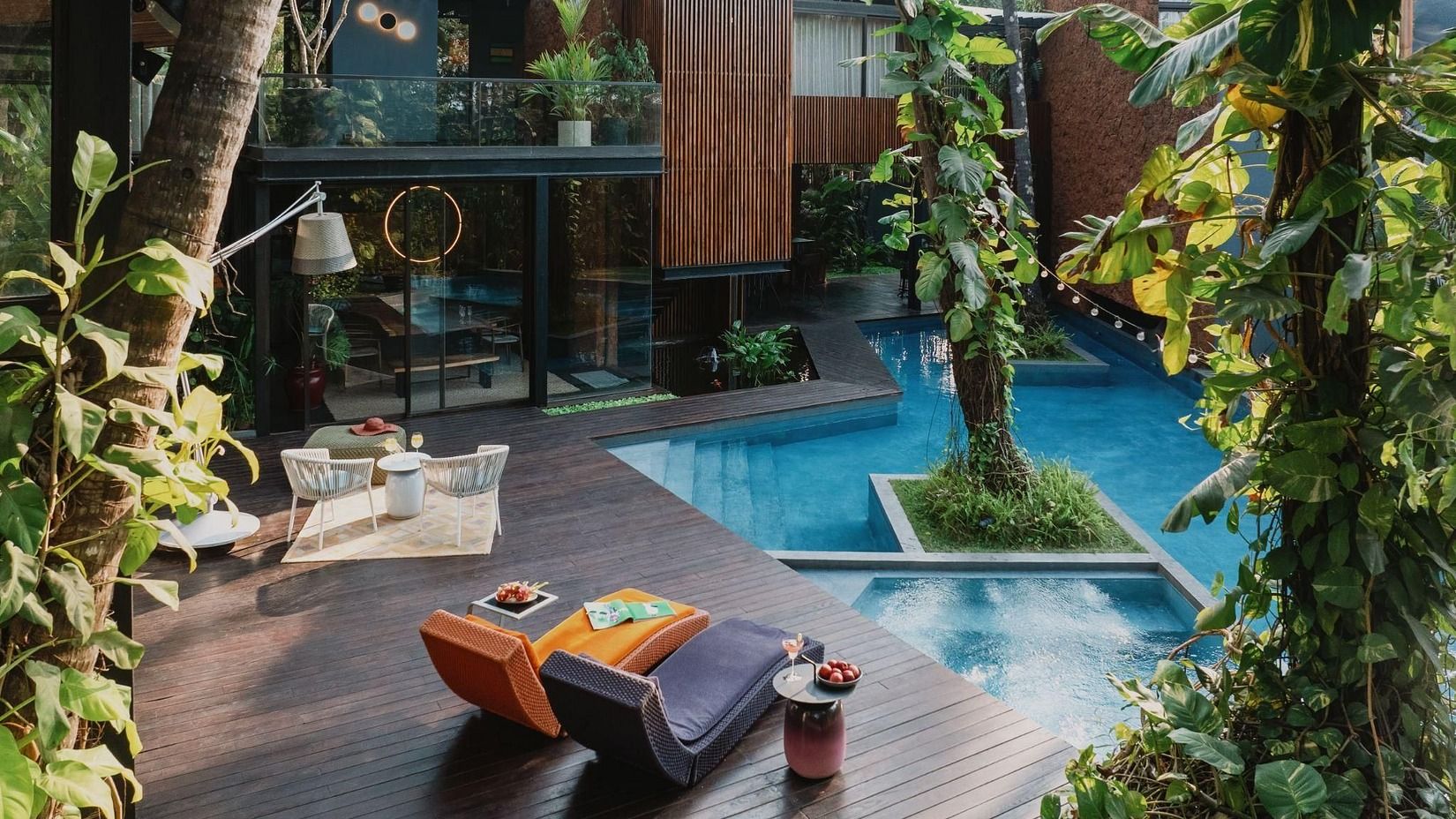 An image of a swimming pool surrounded by plants and on the side of of a pool there are lounger beds