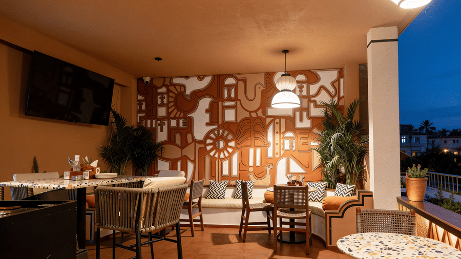 A sheltered outdoor dining patio at The Sierra - By The Lake features wooden tables, chairs, a decorative patterned wall mural, and hanging lamps.