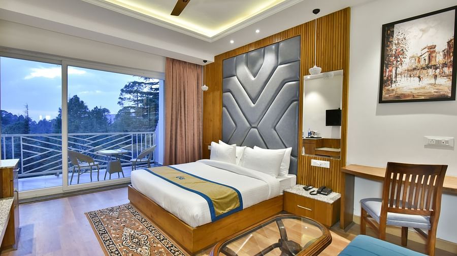 Image of the luxury room with balcony with a bed, bed side tables, chair, work desk, and a glass door leading to the balcony with a scenic view in the background - DLS Ark Holiday Inn, Dalhousie