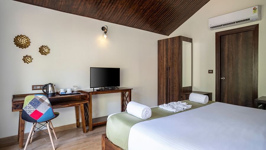 Another view of a hotel bedroom with a large bed with a green runner, a work desk, a colourful chair, a work desk, a wooden cupboard and a wall mounted television - Stone Wood Nature Resort, Gokarna