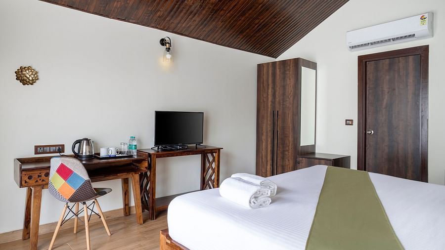 A hotel bedroom with a large bed with a green runner, a work desk, a colourful chair and a wall mounted television - Stone Wood Nature Resort, Gokarna