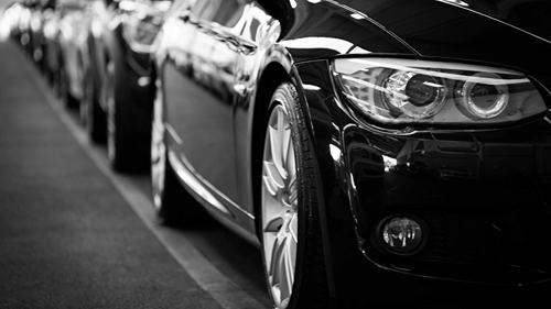 A row of parked cars, lined up along a road with a close-up focus on the front headlight of a black car.