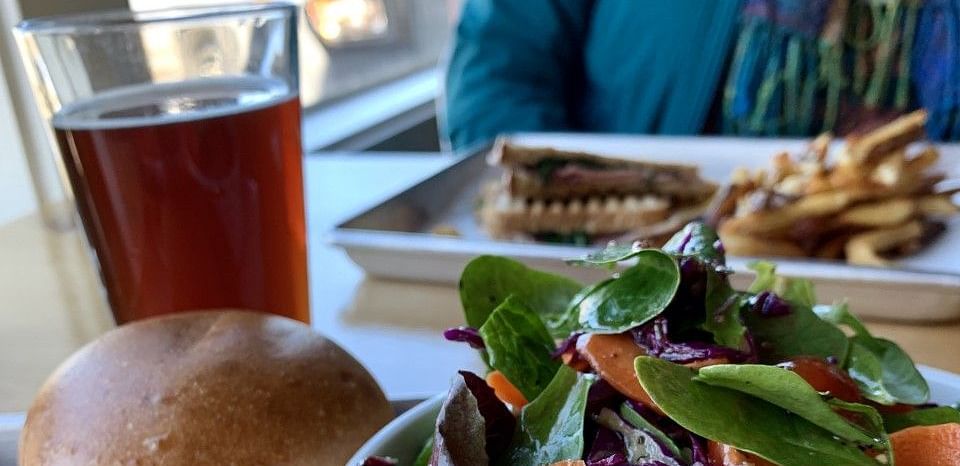 A table with a fresh meal including a burger, a colorful salad, and a glass of beer in a bright dining setting