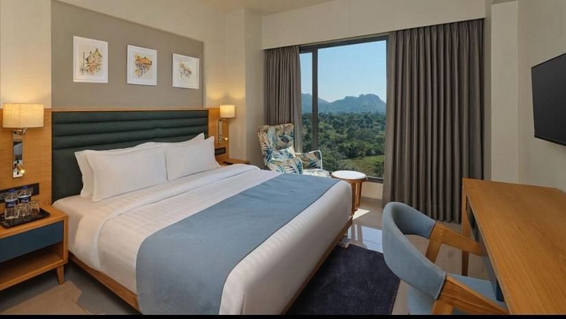 Side view of a room with a double bed and tree frames hanging on a wall and a balcony with scenic view outside an armchair and a tabe at Pride Resort Ambaji