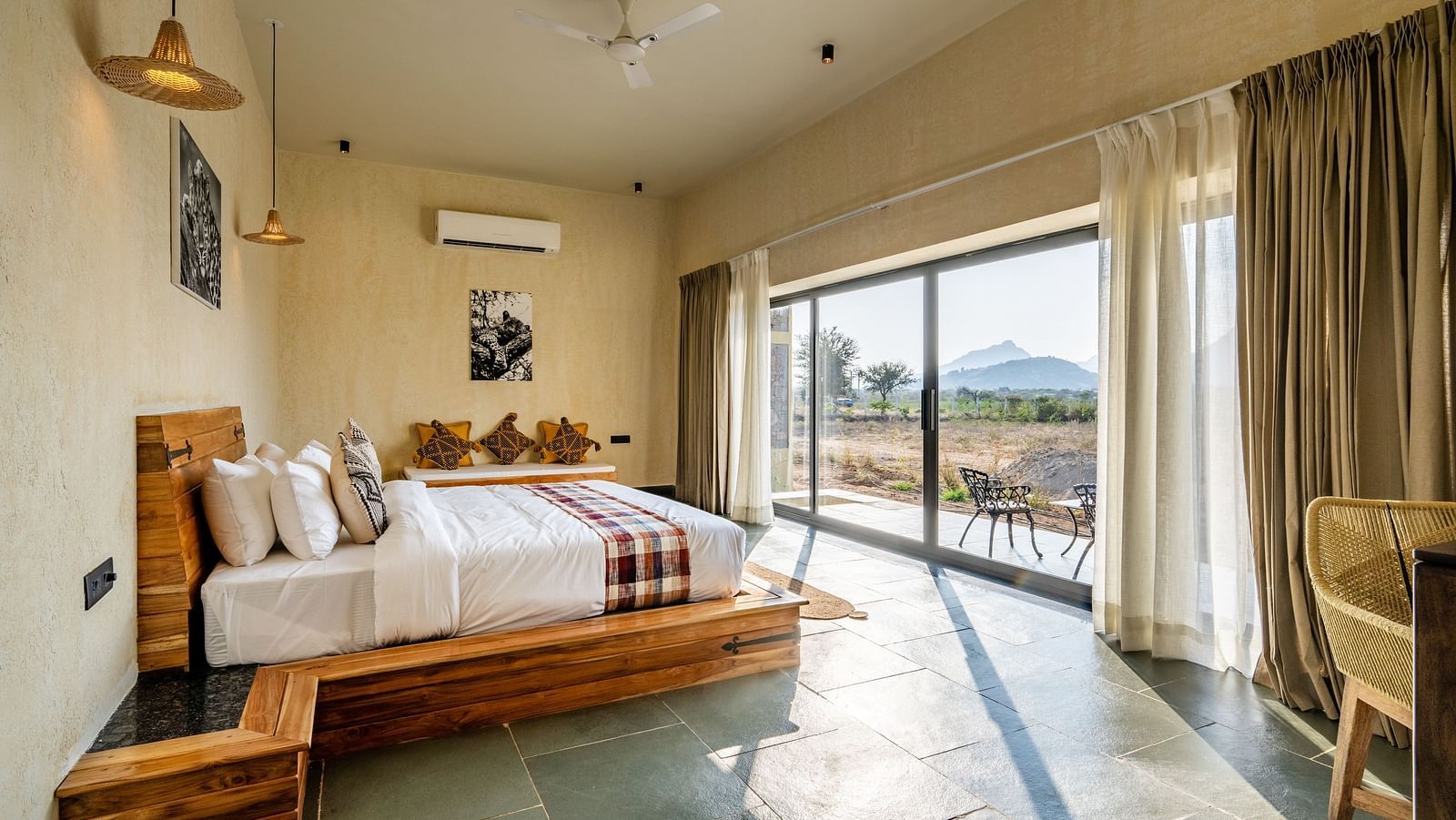  A spacious bedroom with a wooden study table with a chair, a bedside table, a wall-mounted air conditioner, photo frames, and a full-length window view at KURJA Jawai.
