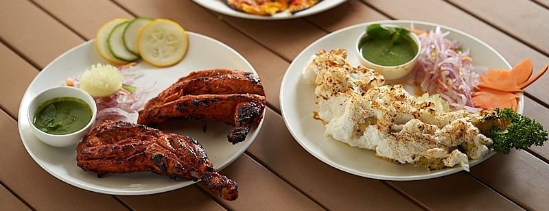 Three plates of food on a wooden table: a sliced vegetable pizza, grilled tandoori chicken with lime and green chutney, and a plate of crispy fried dish with sliced onions and green chutney at The Hamuse Hotel, Kodaikanal.