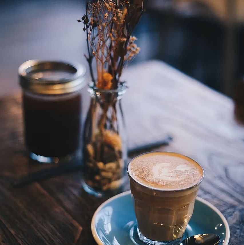 A cup of a coffee served on a blue coaster with a small vase holding dried flowers.