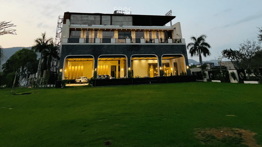 A lawn at Pride Premier Alwar, with an illuminated building, grass, plants, and trees in it.