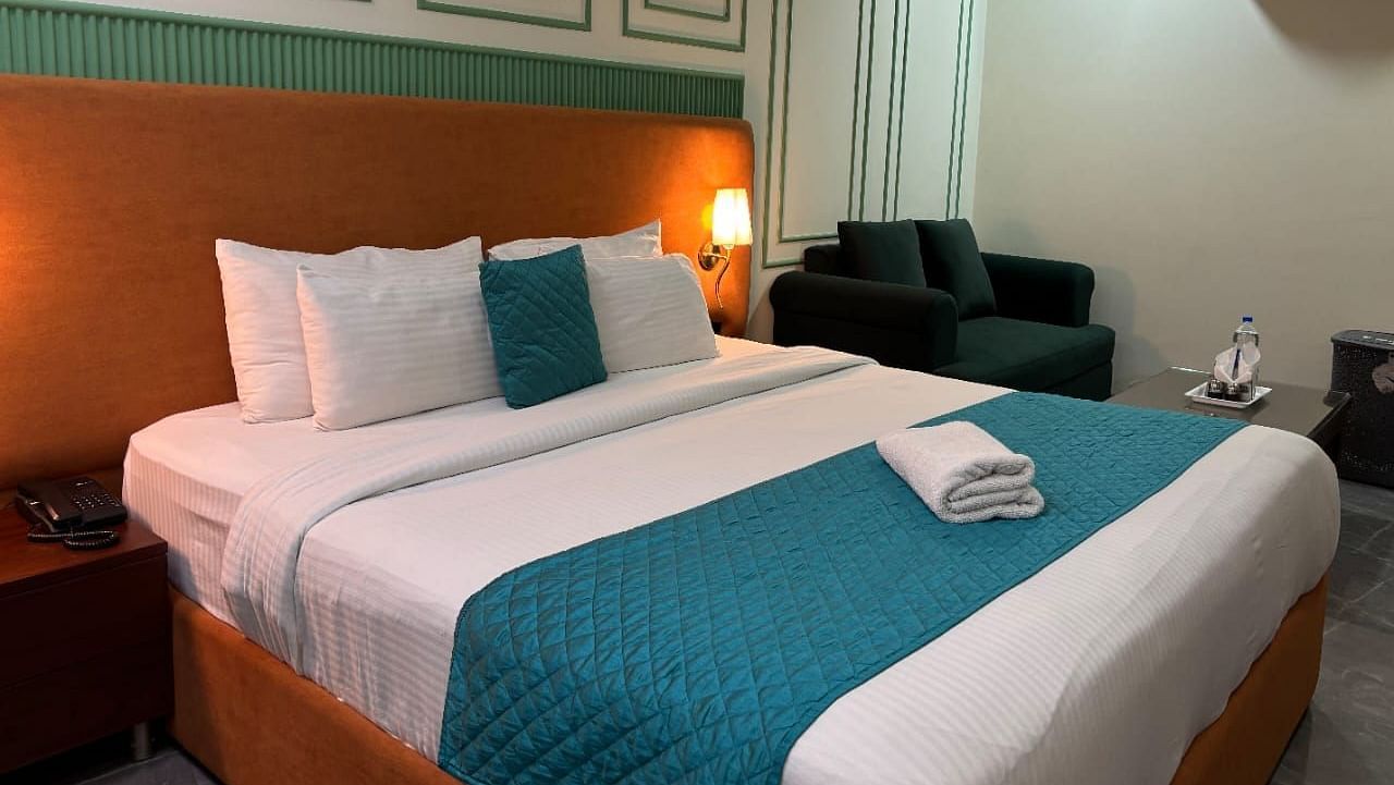 A well-lit room with a double bed dressed in white and blue, and a dark armchair - Asian Suites IFFCO Metro, Gurgaon