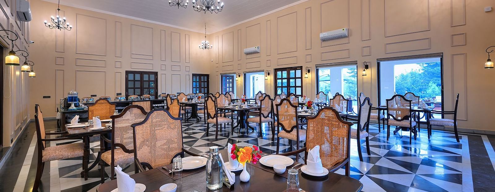 Spacious dining area featuring high ceilings, chandeliers, and classic wooden furniture. The black-and-white patterned flooring contrasts with the warm beige walls, complemented by large windows and elegant wall sconces - Singhvilas, Ranthambore