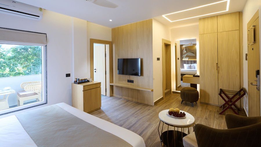 A wide view of a hotel room with a double bed, light wood paneling, a seating area with chairs, and glass doors | Voyage Eastin Suites