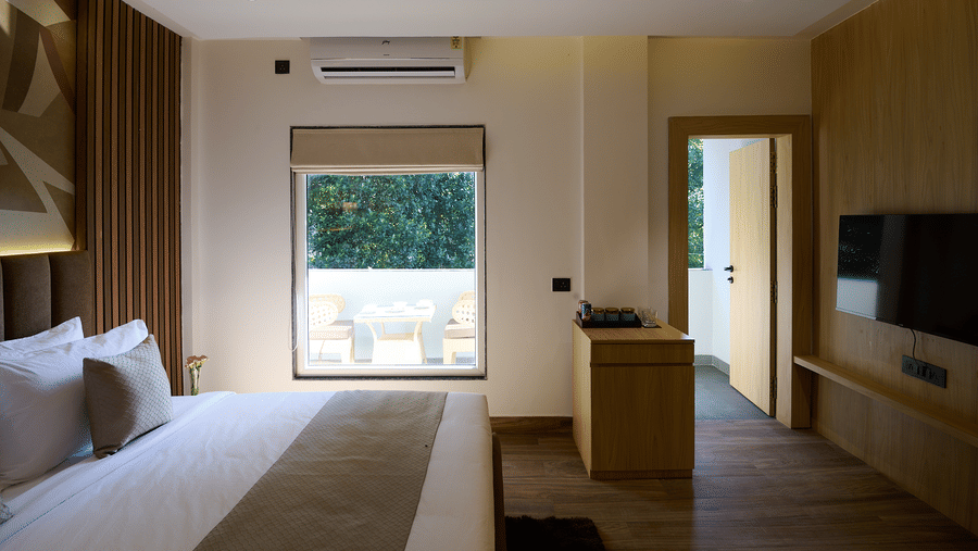 A hotel room with a double bed facing a small balcony or outdoor area visible through the window, and a TV mounted on a wooden wall panel | Voyage Eastin Suites
