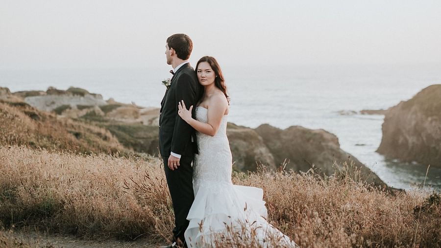 Bride and groom embracing each other on a grassy hill by the ocean