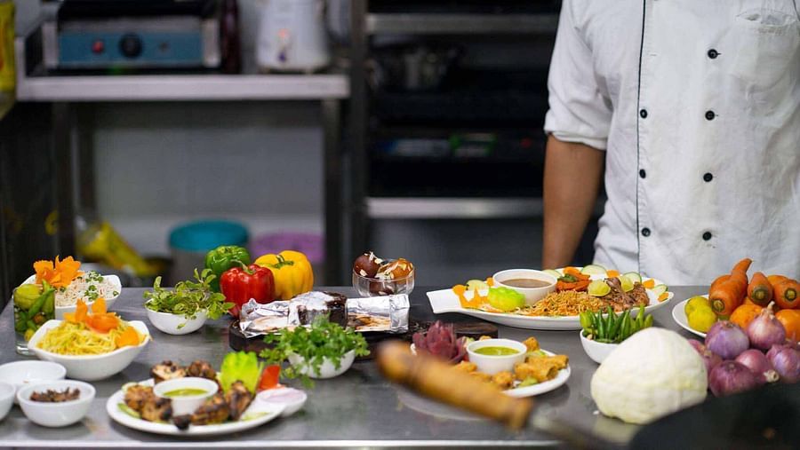 A chef in a white uniform standing behind a counter laden with various ingredients, fresh produce, and prepared dishes at Mansha Hotel Regency.