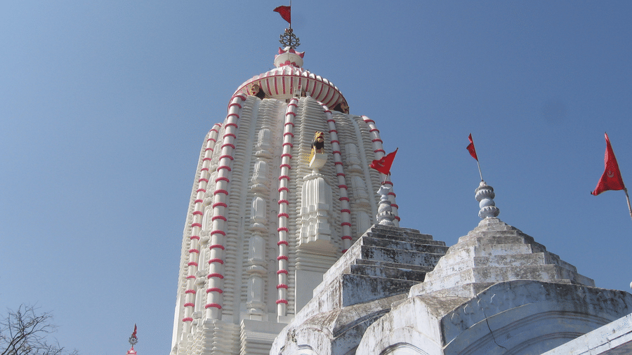 Tall white temple with red vertical stripes, dome top adorned with red flags, smaller domes in foreground against clear blue sky.