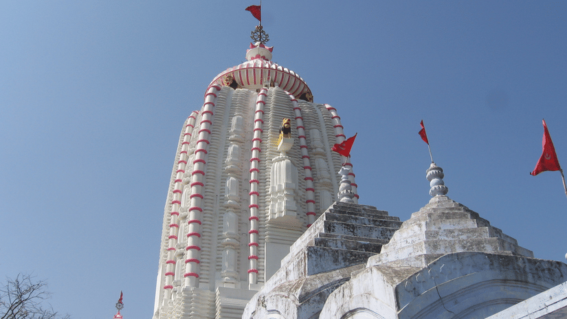 Tall white temple with red vertical stripes, dome top adorned with red flags, smaller domes in foreground against clear blue sky.